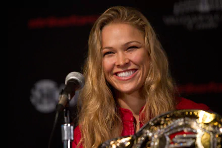 Ronda Rousey smiles confidently at a press conference, holding her championship belt, showcasing her achievements in sports. This HD wallpaper captures her vibrant energy.