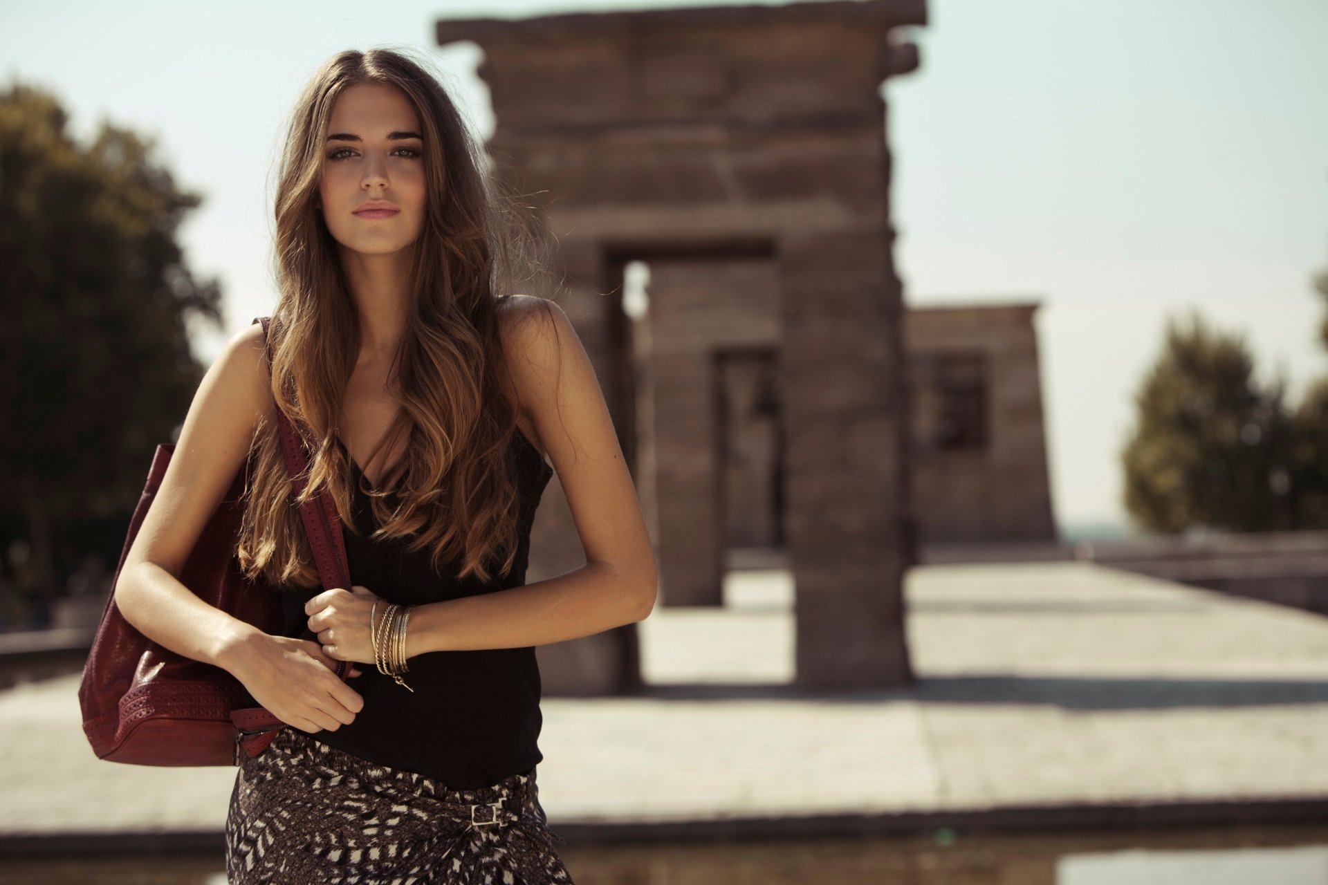 A woman with wavy hair stands confidently in an outdoor setting, holding a bag in her right hand. The scene, tagged for Clara Alonso, serves as a stylish HD desktop wallpaper and background.