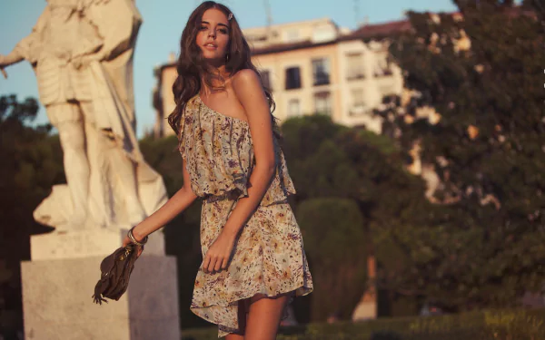 A woman in a floral dress holds a bag, posing outdoors near a statue, with buildings and greenery in the background. This image is tagged with Clara Alonso and is an HD desktop wallpaper and background.