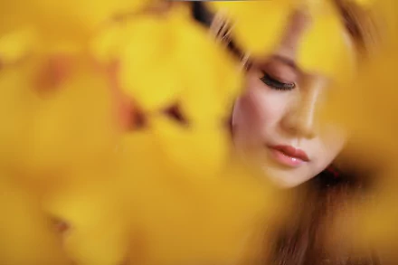 Close-up portrait of a Taiwanese woman partially obscured by vibrant yellow leaves, captured in 4K Ultra HD detail.