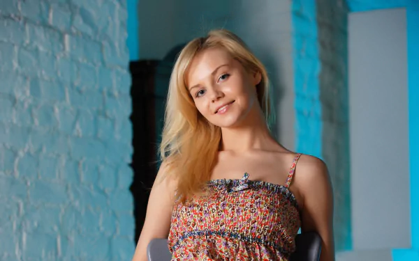 HD desktop wallpaper featuring a beautiful woman with long blonde hair, smiling softly while wearing a patterned top. The background features a white brick wall and modern decor elements.