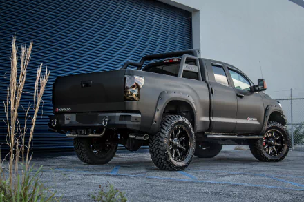 Devolro black Toyota Tundra off-road vehicle with oversized tires and roll bar parked by an industrial garage — 5K Ultra HD PC desktop wallpaper
