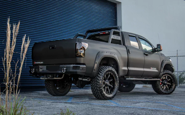 Devolro black Toyota Tundra off-road vehicle with oversized tires and roll bar parked by an industrial garage — 5K Ultra HD PC desktop wallpaper
