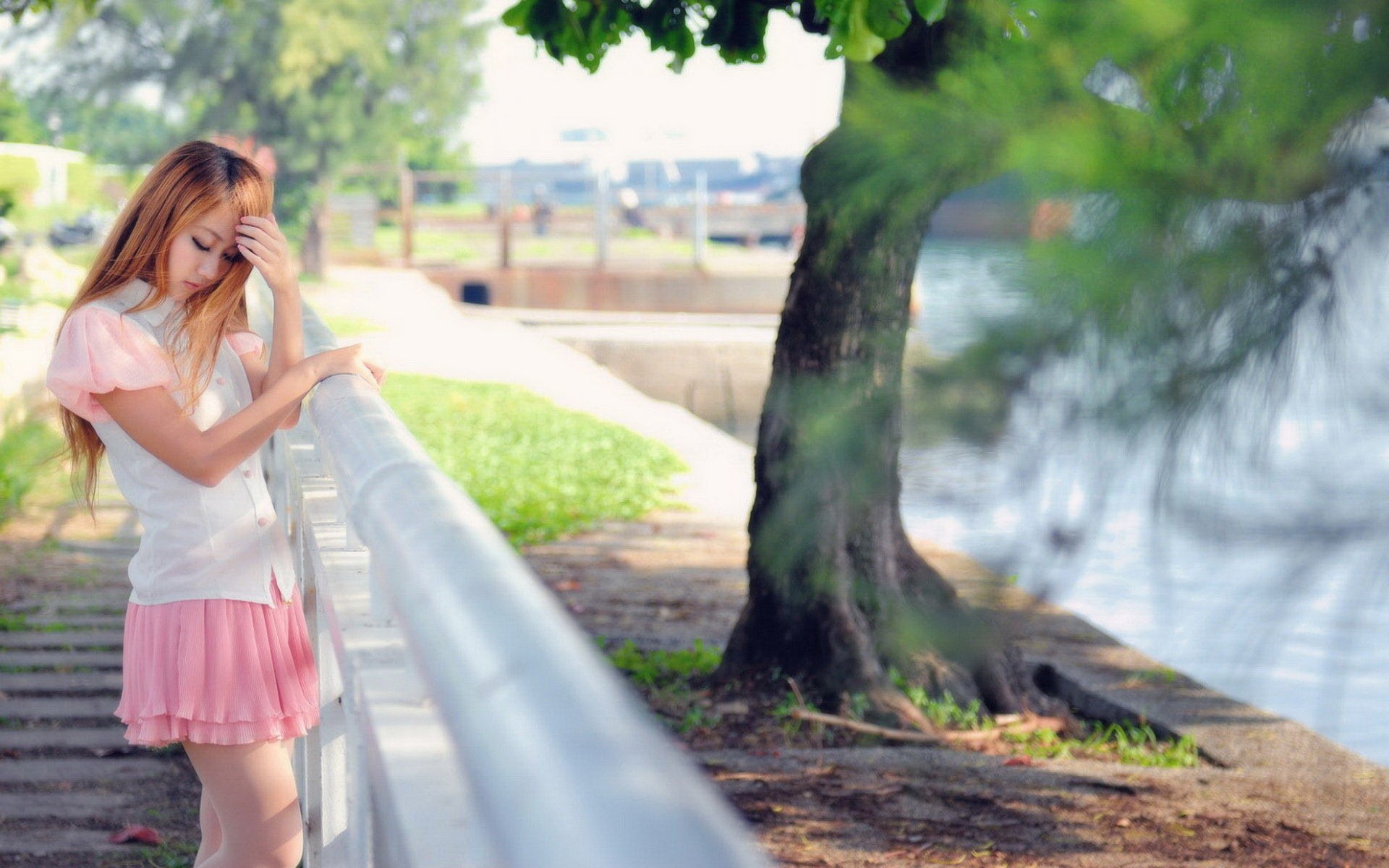 A woman with long, light brown hair stands pensively by a railing near water, framed by lush greenery, creating a serene and contemplative atmosphere. HD desktop wallpaper background.