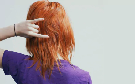 HD music desktop wallpaper of a red-haired musician in a purple shirt, shown from behind making a rock hand gesture against a pale background.