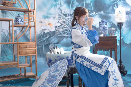 A Taiwanese woman in traditional oriental national dress sips tea indoors, surrounded by blue and white porcelain, ornamental umbrella, and floral decor in a serene room.