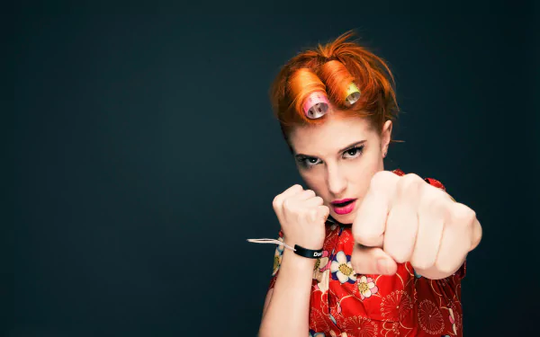 Vibrant 2K Quad HD PC desktop wallpaper: red-haired female musician with curlers and bold makeup throwing a punch toward camera against a deep teal background.
