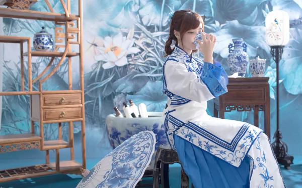 A Taiwanese woman in traditional oriental national dress sips tea indoors, surrounded by blue and white porcelain, ornamental umbrella, and floral decor in a serene room.