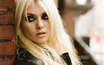2K Quad HD desktop wallpaper of a blonde rock musician with smoky eye makeup and a leather jacket, leaning against a brick wall in an urban, moody portrait.