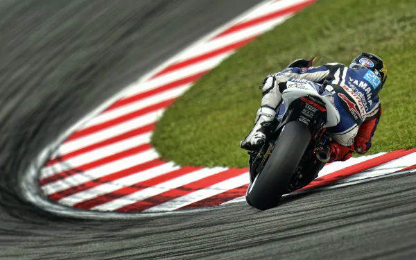 HD PC desktop wallpaper/background: Yamaha racing vehicle leaning into a curving racetrack, rider in blue-white leathers, motion-blurred asphalt and red-and-white curb.