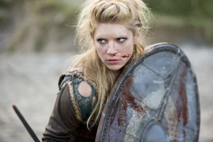 A fierce Katheryn Winnick as Lagertha from the TV show Vikings (2013), stands ready for battle, wielding a shield with a determined gaze and signs of a recent fight.