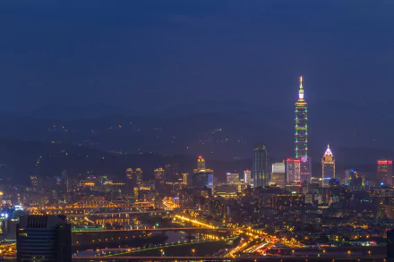 Nighttime view of Taipei cityscape featuring the illuminated Taipei 101 skyscraper, surrounding buildings, and distant mountains under a clear dark sky.