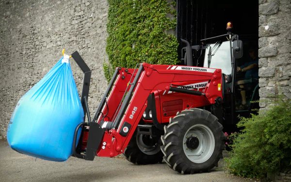 A Massey Ferguson tractor is featured lifting a large blue bag, parked at the entrance of a stone structure, creating a striking HD desktop wallpaper.
