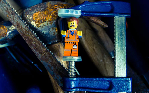 HD PC desktop wallpaper featuring Emmet from The Lego Movie, a man-made Lego figure, clamped tightly in a metal vise against a dark, textured background.