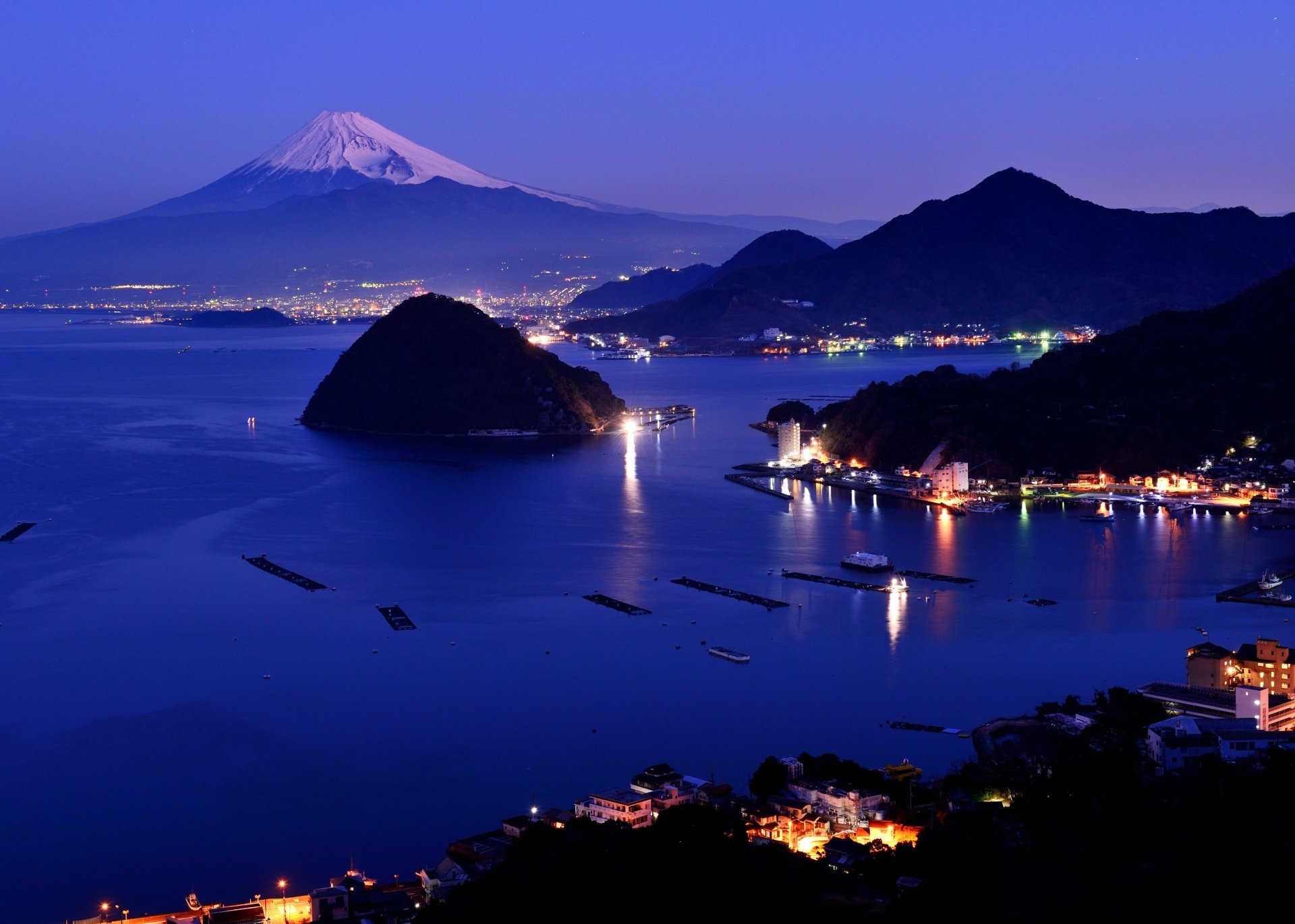 4K Ultra HD night landscape of Mount Fuji, an iconic volcano in Japan, overlooking a calm coastal area illuminated by city lights and surrounded by dark hills.