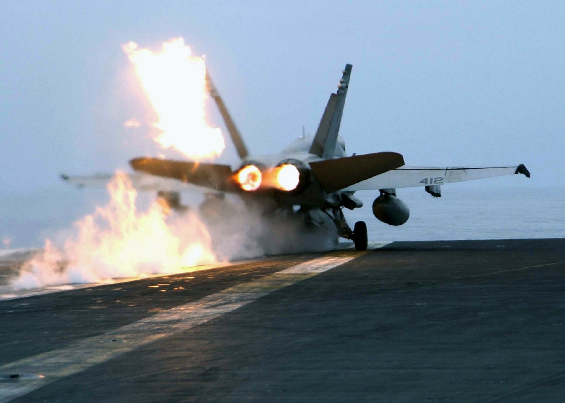 HD PC desktop wallpaper of a military McDonnell Douglas F/A-18 Hornet launching from an aircraft carrier, afterburners blazing as flames flare on the flight deck.