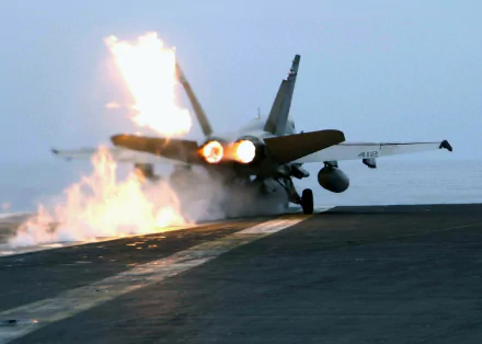 HD PC desktop wallpaper of a military McDonnell Douglas F/A-18 Hornet launching from an aircraft carrier, afterburners blazing as flames flare on the flight deck.