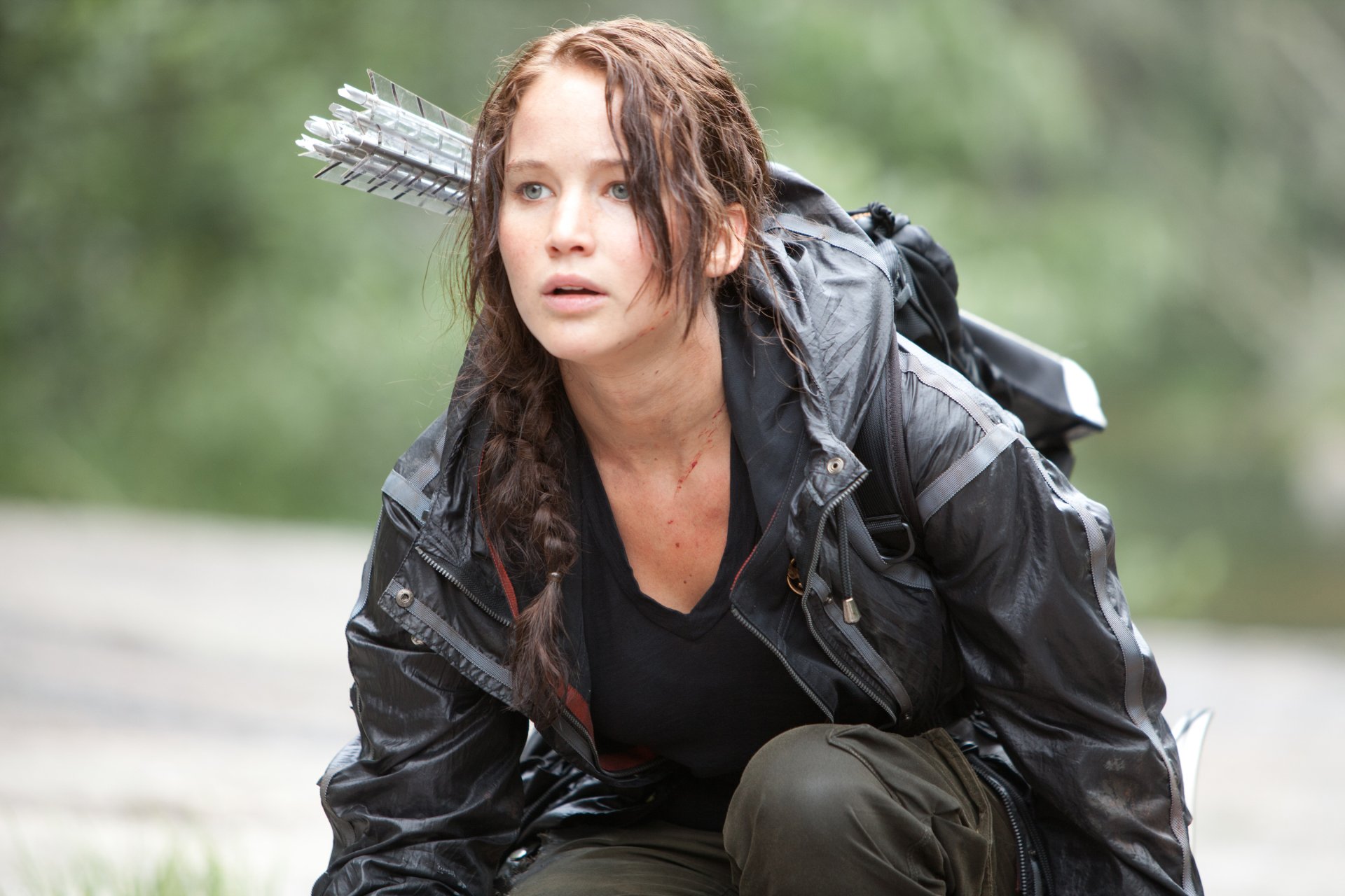 Crouching woman in a wet dark jacket with arrows on her back in a forested movie scene — The Hunger Games 2K Quad HD PC wallpaper/background