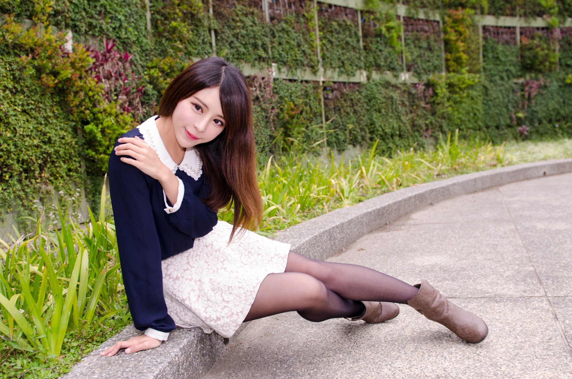 A smiling Taiwanese model, Julie Chang, poses outdoors in a white dress and black sweater, set against a lush green wall, captured in 4K Ultra HD quality.