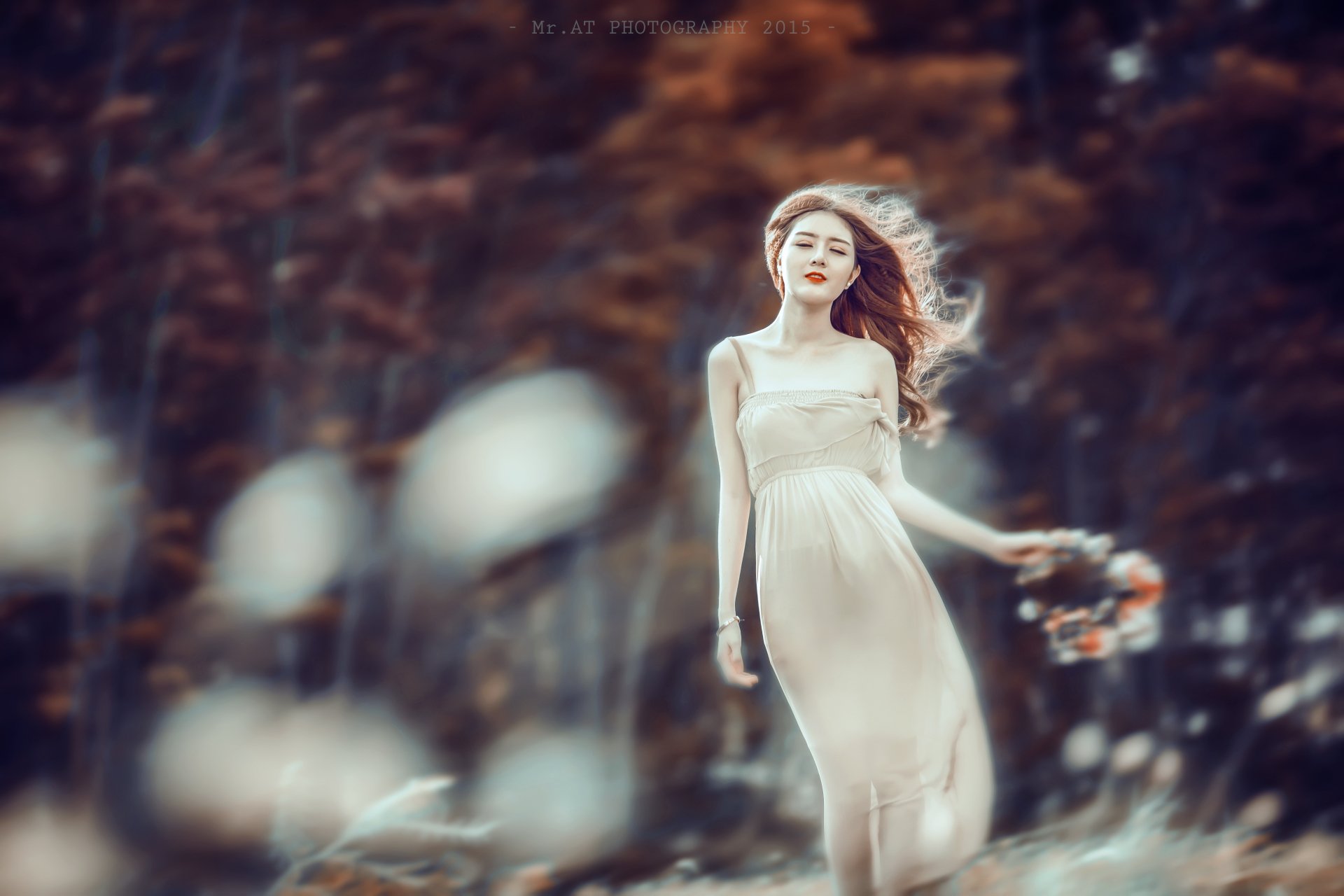A Vietnamese woman in a flowing dress stands gracefully outdoors, surrounded by soft bokeh and autumnal tones, wearing a delicate wreath in her hair.