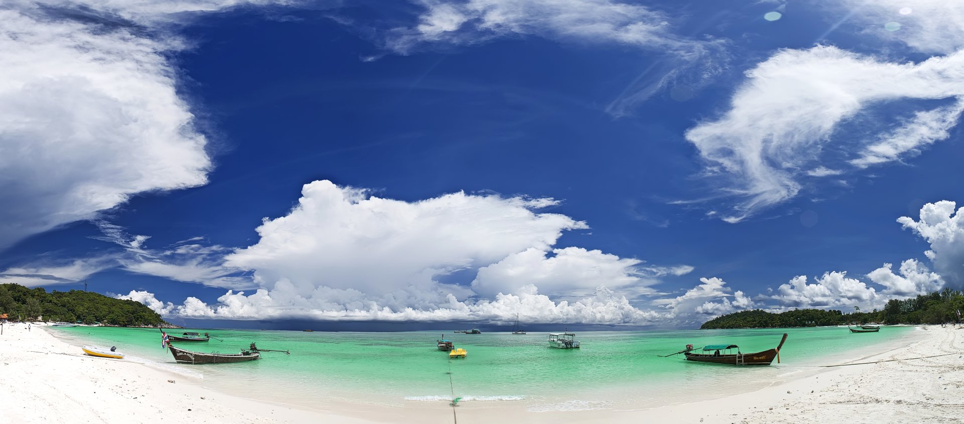 A panoramic view of a tropical beach in Thailand, featuring clear blue waters, white sand, and boats anchored in a tranquil lagoon under a vibrant sky with fluffy clouds.