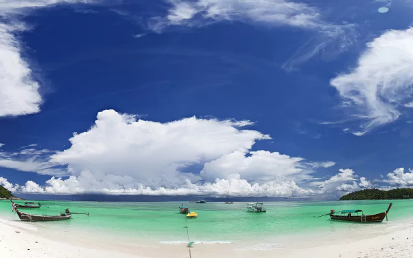 A panoramic view of a tropical beach in Thailand, featuring clear blue waters, white sand, and boats anchored in a tranquil lagoon under a vibrant sky with fluffy clouds.