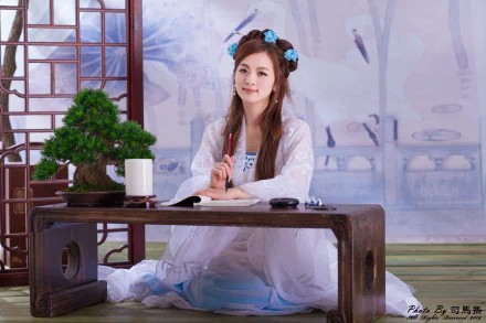 A smiling Asian woman dressed in a traditional Chinese costume sits at a table with a bonsai and brush, showcasing elegance and cultural beauty. This 4K Ultra HD image captures a serene moment.