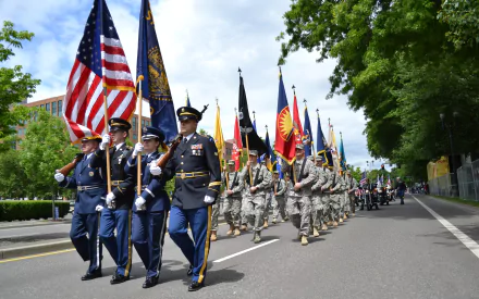 HD PC desktop wallpaper showing a Memorial Day parade with uniformed military members marching and carrying American flags along a tree-lined street.