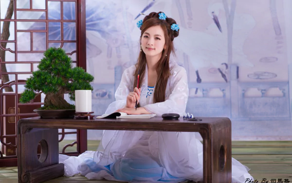 A smiling Asian woman dressed in a traditional Chinese costume sits at a table with a bonsai and brush, showcasing elegance and cultural beauty. This 4K Ultra HD image captures a serene moment.