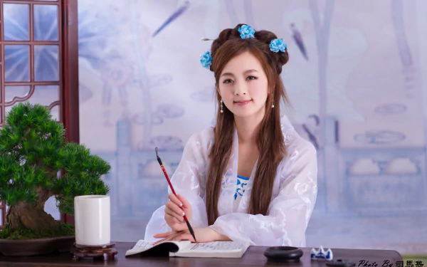 Young woman in traditional costume with hairpins and blue floral hair ornaments, holding a brush over an open book beside a bonsai on a desk — 4K desktop wallpaper.