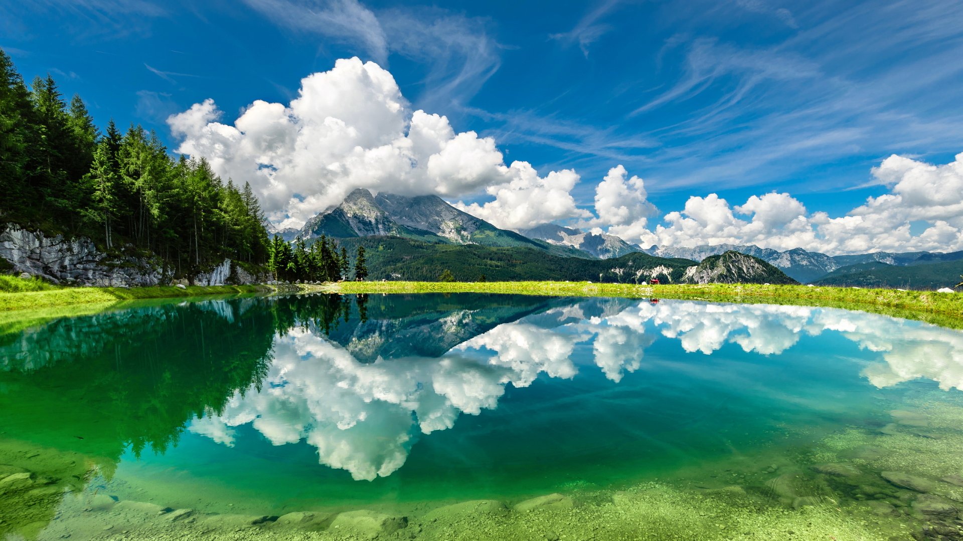 HD desktop wallpaper featuring a breathtaking nature scene with a clear mountain lake reflecting fluffy white clouds and a vivid blue sky.