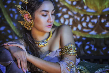 A Thai woman with elegant jewelry, including a necklace and earrings, poses gracefully in front of ornate background decor, showcasing traditional beauty in a captivating HD image.