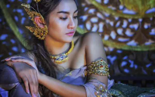 A Thai woman with elegant jewelry, including a necklace and earrings, poses gracefully in front of ornate background decor, showcasing traditional beauty in a captivating HD image.