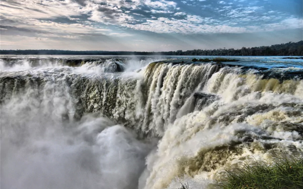 A stunning view of Iguazu Falls, showcasing the powerful waterfalls straddling the border of Argentina and Brazil, surrounded by lush nature under a partly cloudy sky.