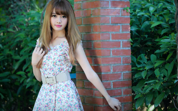 A Taiwanese model, Cubie Wang, stands gracefully by a brick wall, wearing a floral dress cinched with a stylish belt, surrounded by lush green foliage. 
