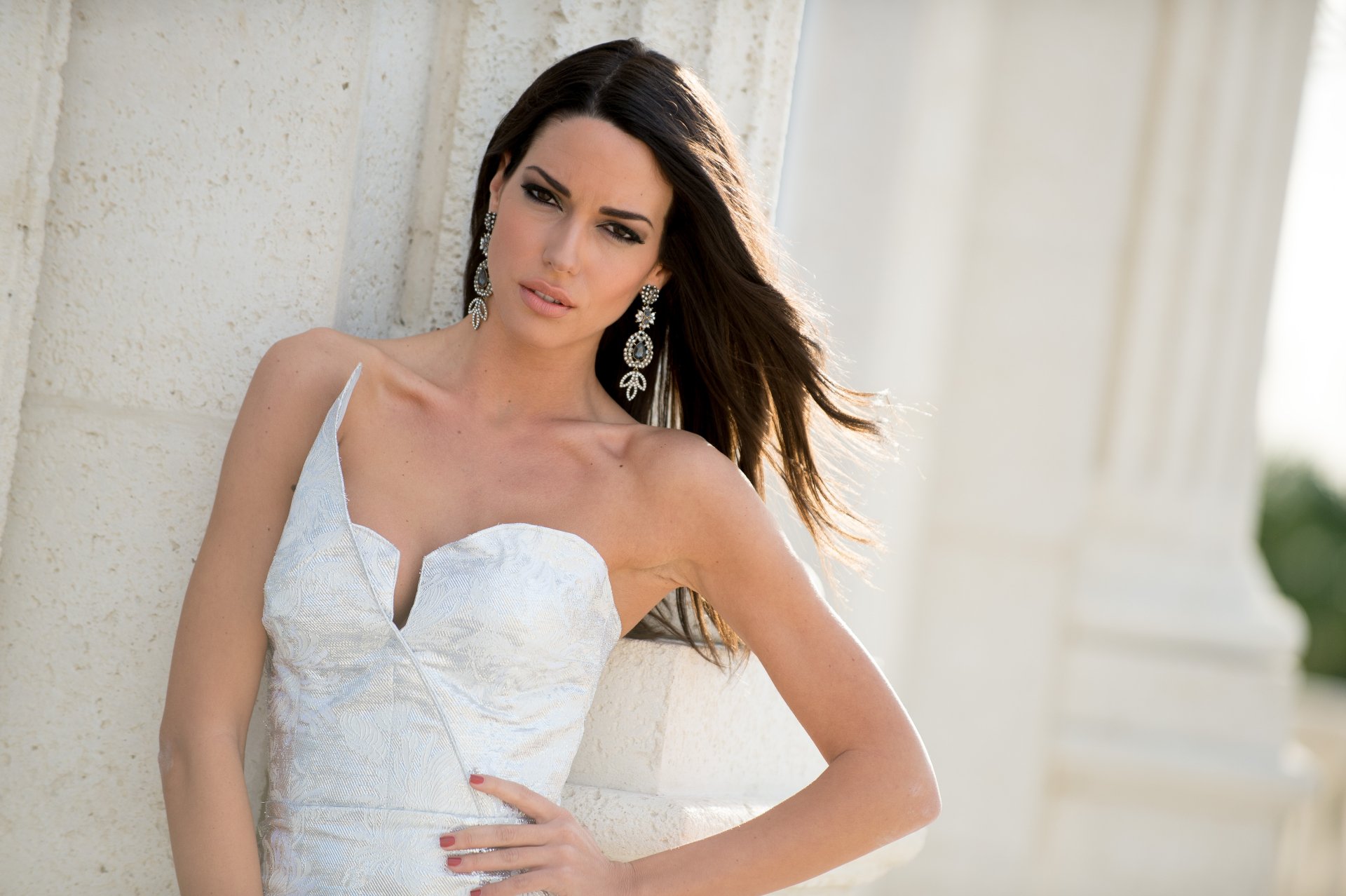 Greek model Ismini Dafopoulou, Miss Greece, poses in a white dress and elegant earrings against a sunlit stone backdrop in this 4K Ultra HD desktop wallpaper.