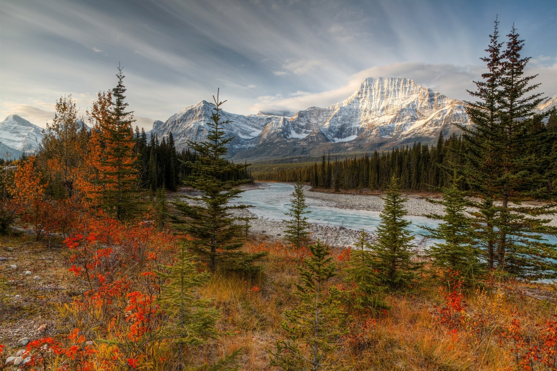 HD desktop wallpaper showcasing a vibrant forest with autumn foliage beside a winding river, set against towering mountain peaks under a cloudy sky in a stunning natural landscape.