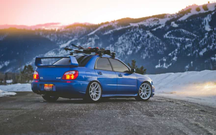 A blue Subaru Impreza WRX parked on a mountain road with snow-covered peaks and a sunset sky in the background, captured in high-definition for a desktop wallpaper.