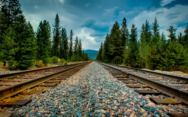 A serene view of a railroad track stretching into the distance, surrounded by lush greenery and mountains under a dramatic sky, making a striking HD desktop wallpaper.