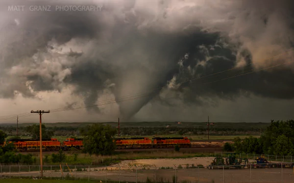 A powerful tornado churns over a Texas railroad track as a train moves through the storm under dark, ominous clouds in this HD desktop wallpaper capturing nature’s fury.