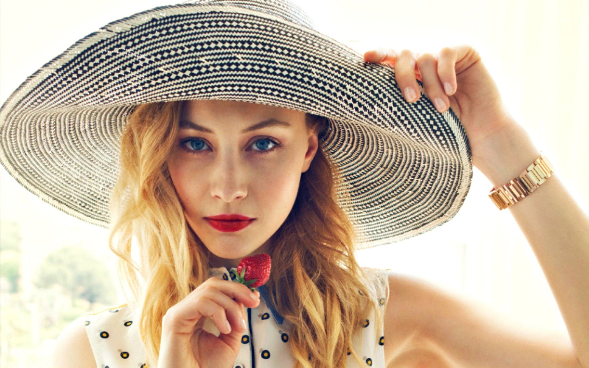 Close-up of Canadian actress Sarah Gadon with blue eyes and blonde hair, wearing a wide-brimmed hat and holding a small strawberry, against a bright background.