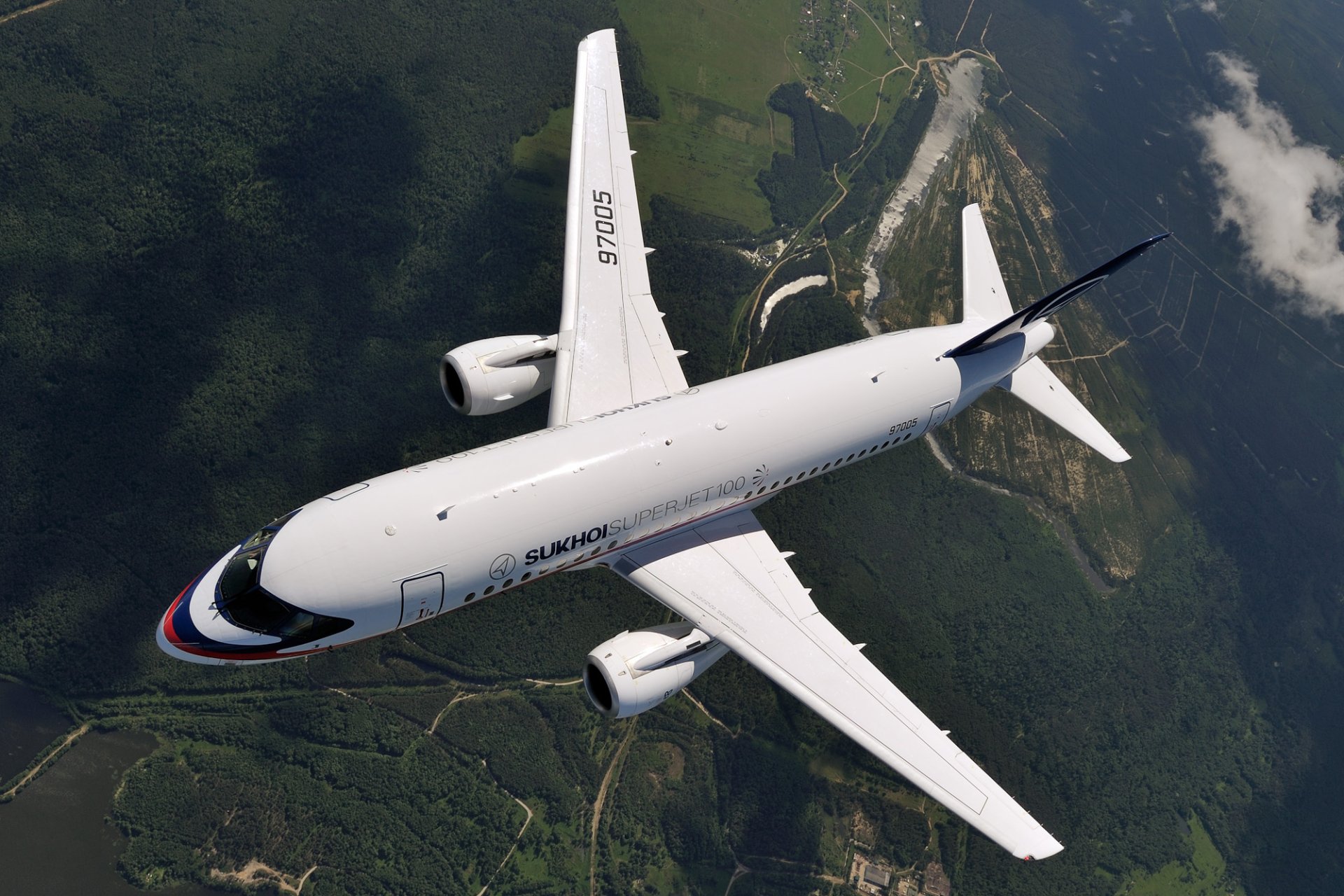 HD PC desktop wallpaper of a Sukhoi SuperJet 100 jet airplane vehicle in flight, top-down view over green terrain and a winding river beneath a dramatic sky.