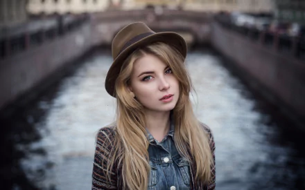 A blonde woman with blue eyes wears a stylish hat, posing against a backdrop of flowing water and a bridge, creating a striking HD desktop wallpaper.
