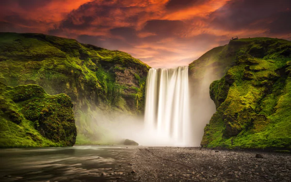 Skógafoss waterfall in Iceland glowing under a vibrant sunset sky, surrounded by lush green cliffs and mist rising from the water, captured in HD for a desktop wallpaper.