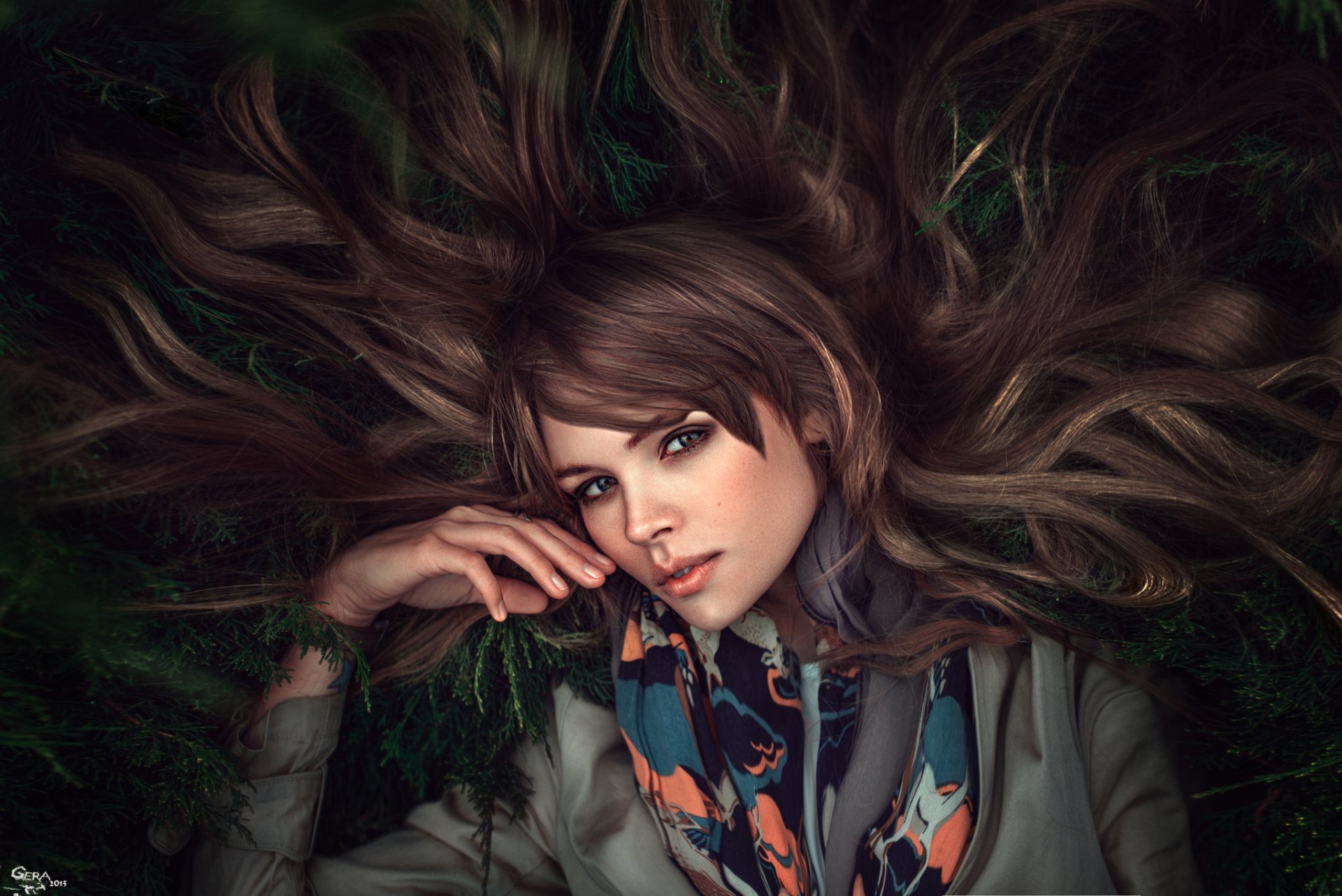 A brunette model with striking green eyes rests amid flowing hair and greenery, showcasing a serene expression. This HD image captures a natural and captivating beauty.