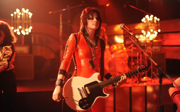Kristen Stewart performs in a vibrant scene from the movie The Runaways, dressed in a striking orange outfit and holding a guitar, capturing the energy of the band's live performance.