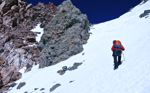 4K Ultra HD PC wallpaper: mountaineer in red jacket climbs a stone-strewn snowy mountain slope under a deep blue sky — winter nature, snow sports scene.