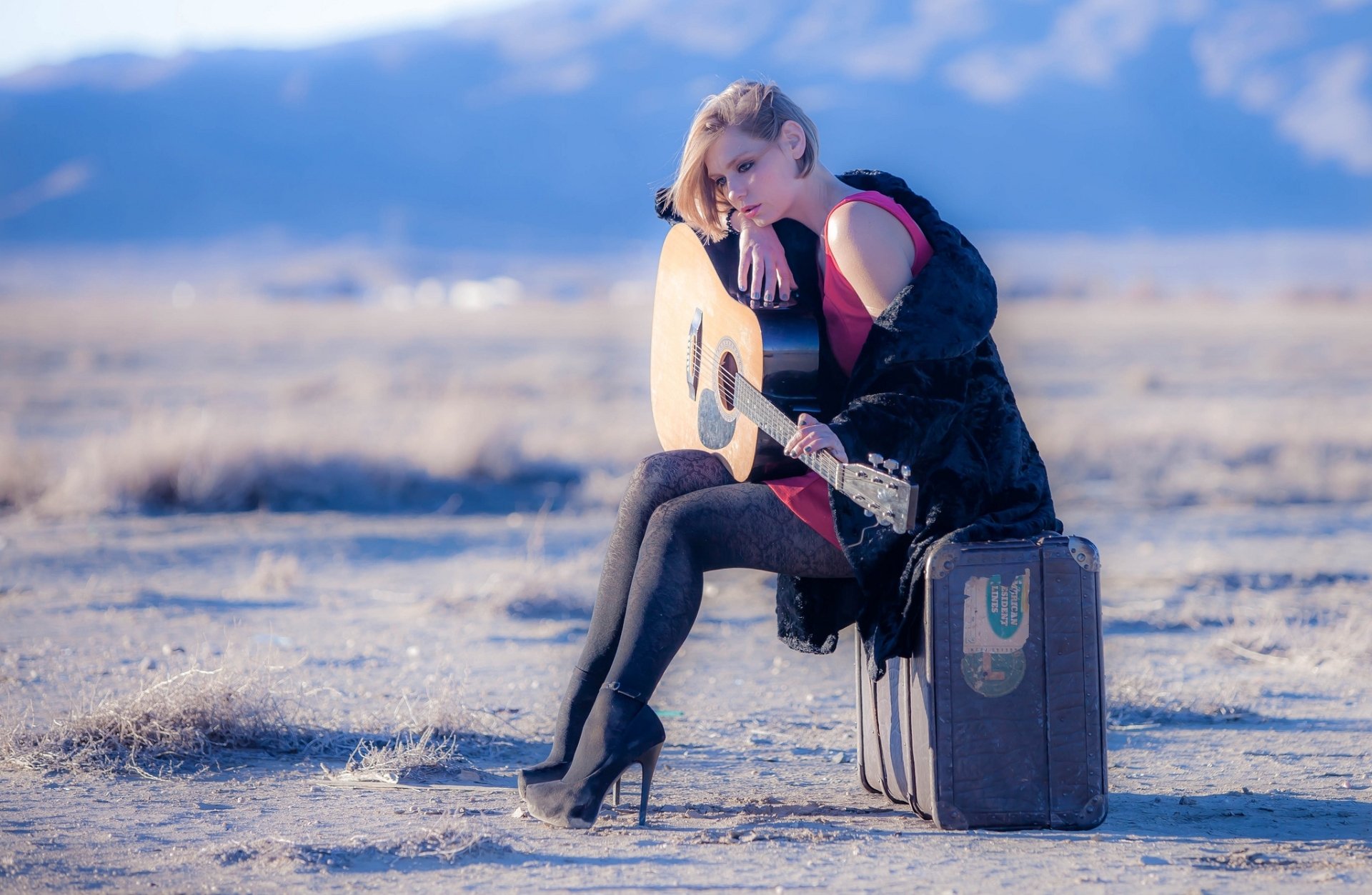 HD PC desktop wallpaper featuring a woman in a contemplative mood, sitting outdoors with a guitar on a vintage suitcase against a serene landscape backdrop.