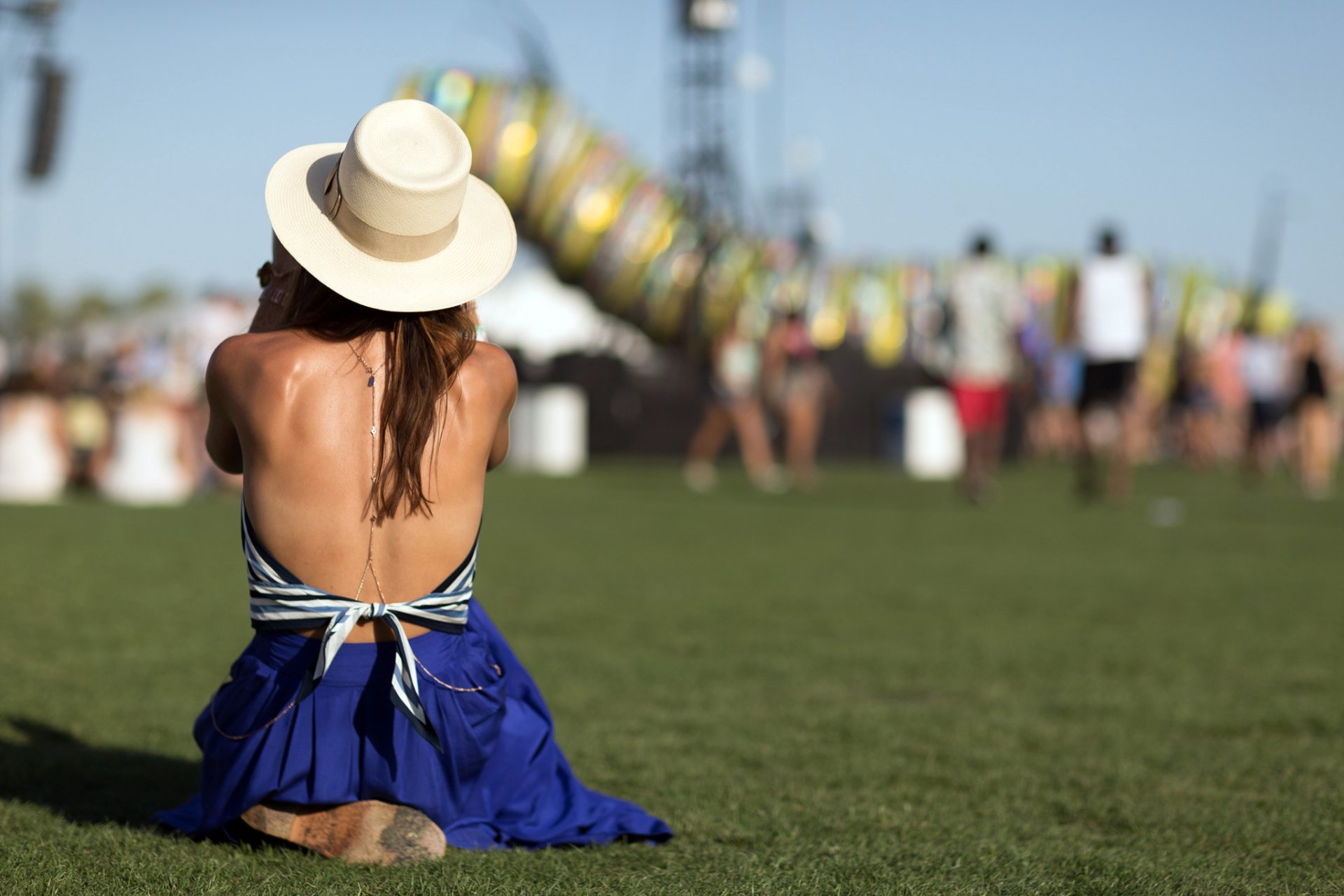 HD PC desktop wallpaper: rear view of a woman in a blue dress and sunhat, sitting on grass at an outdoor festival with blurred crowd and rides in the background.