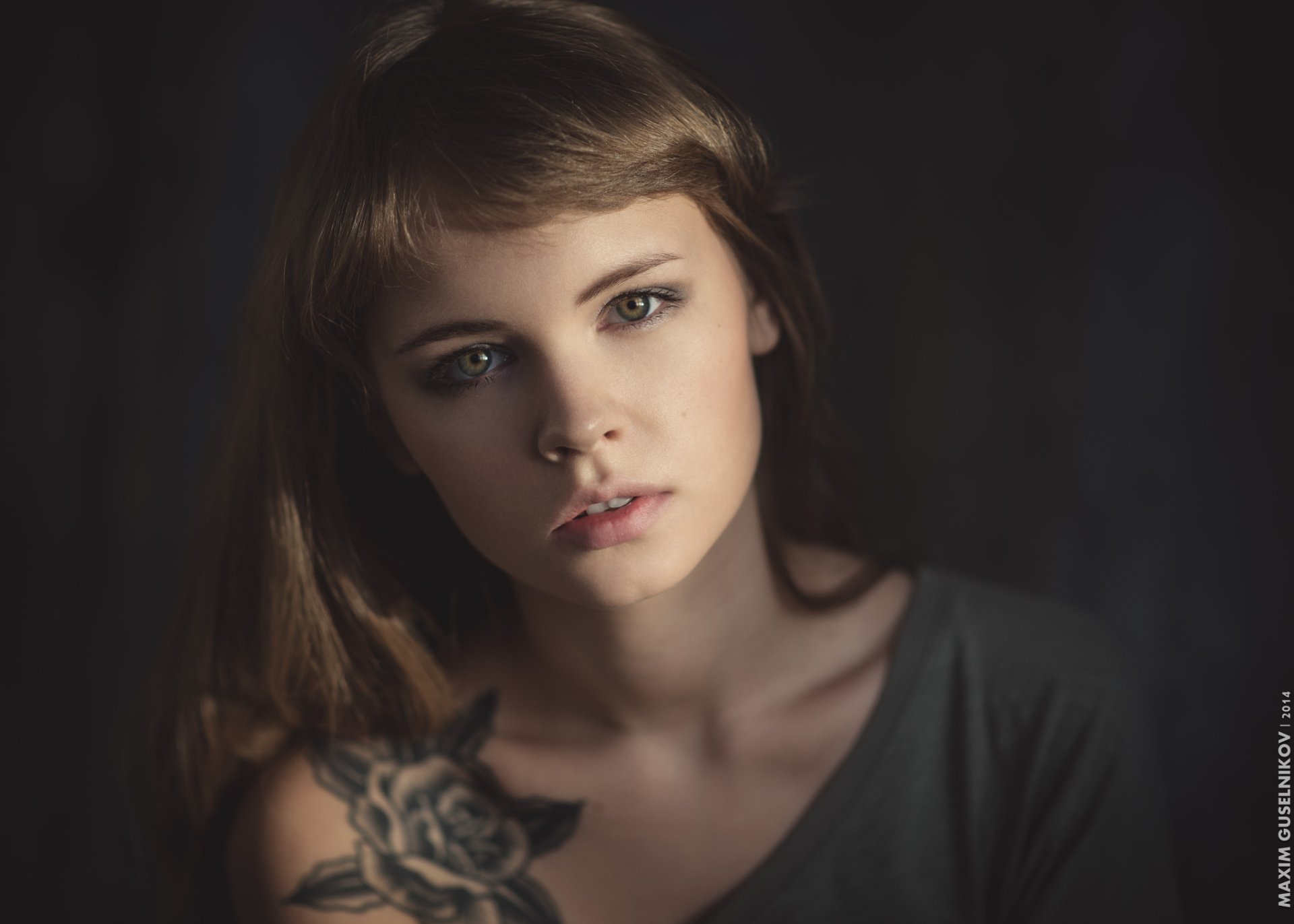 A close-up of a brunette woman with striking green eyes and a visible tattoo, showcasing her intense expression in soft lighting. This HD wallpaper features Anastasiya Scheglova.